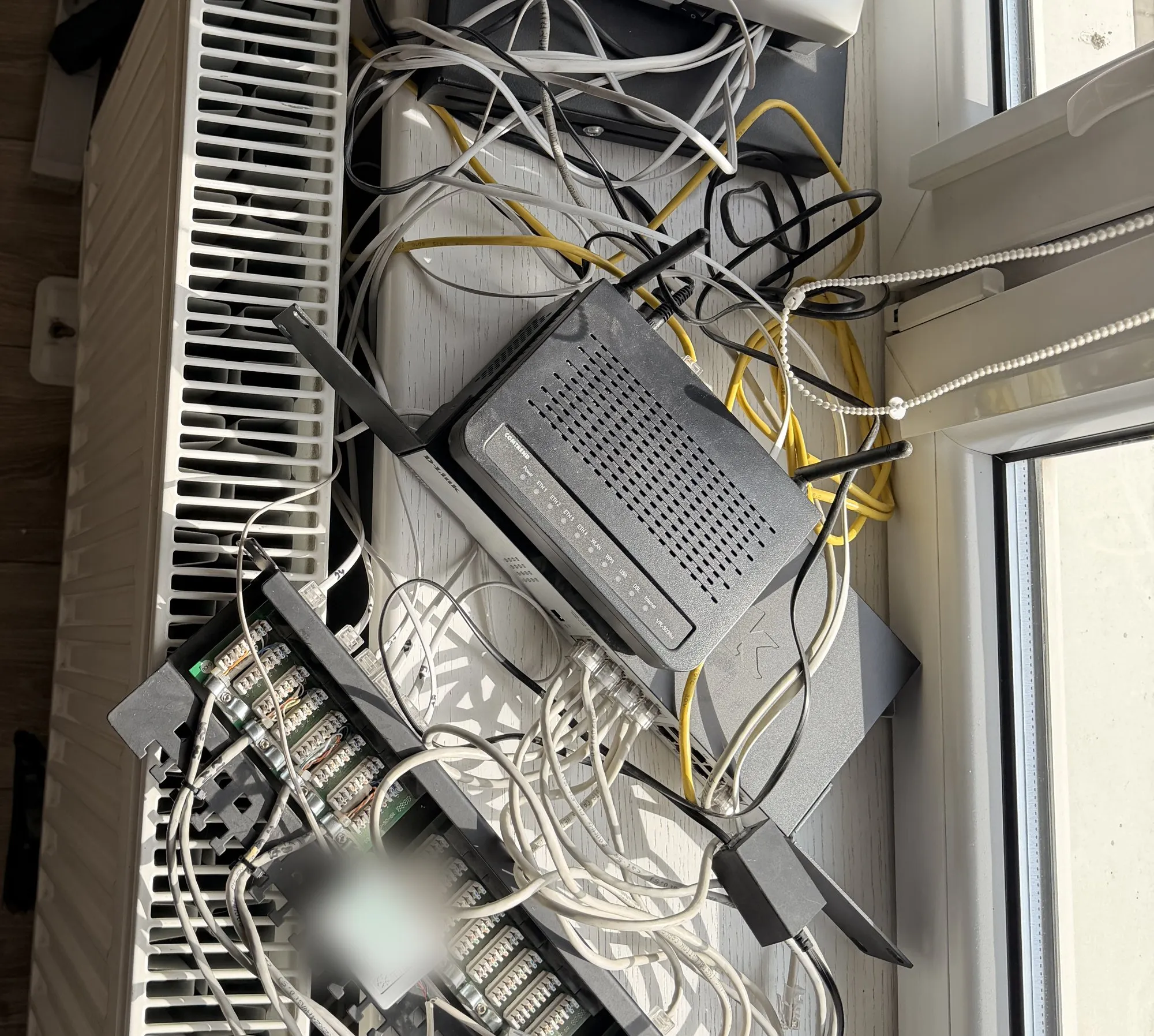 Router, przełącznik, centrala telefoniczna i splątane przewody ułożone na parapecie nad grzejnikiem